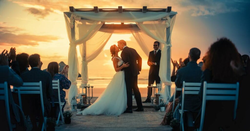 A young bride and groom kissing while celebrating their beach wedding ceremony in front of friends and family.